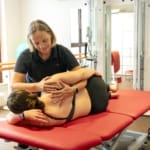 Ein Physiotherapeut hilft einer Frau bei Dehnungsübungen auf einem rot gepolsterten Tisch in einem Fitnessstudio.