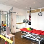 Ein Physiotherapieraum mit Trainingsgeräten, einer roten Behandlungsliege, Balancebällen, Seilen und einer Wanduhr.