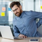 Ein Mann mit Brille und blauem Hemd sitzt mit einem Laptop an einem Schreibtisch und hält sich dabei den unteren Rücken. An der Glaswand hinter ihm hängen bunte Haftnotizen, auf dem Schreibtisch liegt ein Smartphone.