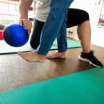 Person in Trainingskleidung balanciert mit einem blauen Ball auf einem Bein auf einer grünen Matte und wird dabei von einem Trainer unterstützt.