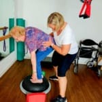 Eine ältere Frau balanciert mit Unterstützung eines Trainers in einem Fitnessraum auf einer Stabilitätsplattform. Ein Rollator und Trainingsgeräte stehen in der Nähe.