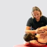 Eine Frau in Sportkleidung liegt auf einem roten Massagetisch, während eine professionelle Massagetherapeutin im schwarzen Hemd eine therapeutische Massage an ihrem Rücken und ihren Schultern durchführt.