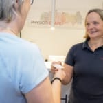 Eine Frau überreicht einer anderen Frau im Empfangsbereich einer Physio-Praxis eine Visitenkarte.