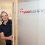 Eine Frau im dunklen Poloshirt steht in einer Praxis vor einer Tür mit der Aufschrift „PhysioGEHRKEN Physiotherapiepraxis“.