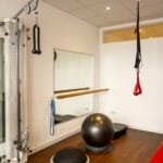 Ein Fitnessraum mit Trainingsgeräten, darunter eine Kabelmaschine, ein Spiegel, Balancebälle, ein Übungsband, ein TRX-Gurt und eine rote Bank auf Holzboden.