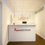 Ein moderner Empfangsbereich für Physiotherapie mit einem weißen Schreibtisch, Branding auf der Vorderseite, einem Stuhl im Hintergrund und Trainingsgeräten an der Seite.