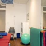 Physiotherapieraum mit Behandlungstisch, Gymnastikball, Widerstandsbändern, Stuhl und offenen Türen.