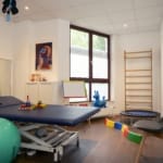 Ein Therapieraum mit Behandlungstisch, Gymnastikball, Spielzeug, Trampolin, Kletterleiter und Stuhl. Die Wände sind mit abstrakter Kunst dekoriert. Ein Fenster sorgt für Tageslicht.