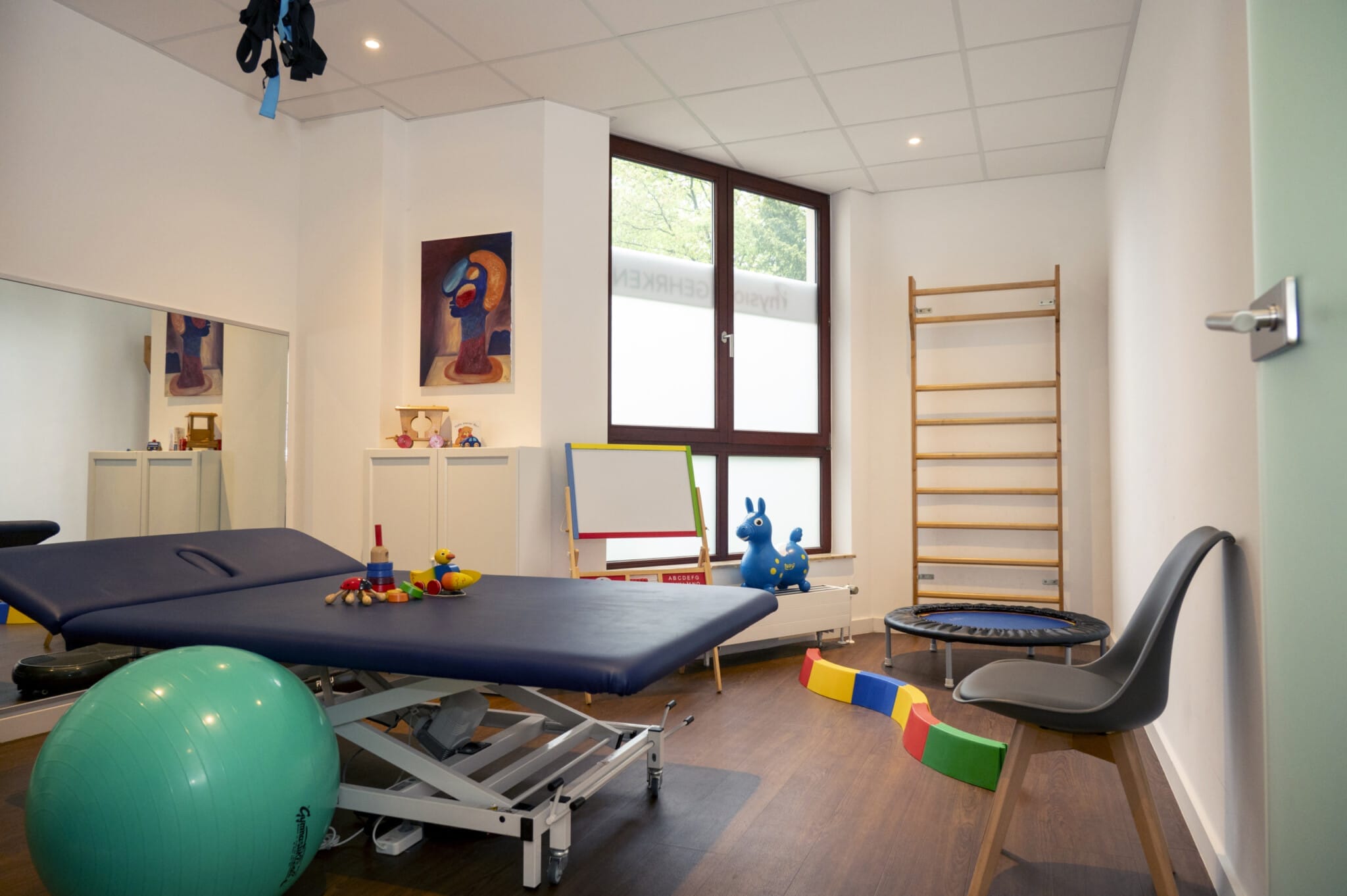 Ein Therapieraum mit Behandlungstisch, Gymnastikball, Spielzeug, Trampolin, Kletterleiter und Stuhl. Die Wände sind mit abstrakter Kunst dekoriert. Ein Fenster sorgt für Tageslicht.