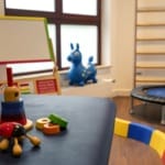 Ein Kinderspielzimmer mit stapelbarem Spielzeug auf einem Tisch, einer Staffelei, einem Trampolin, einem Schaukelpferd und Klettergerüsten.