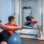 Ein Kind im roten Hemd balanciert auf einem blauen Gymnastikball, während eine Frau ihm hilft; beide spiegeln sich in einem Spiegel.