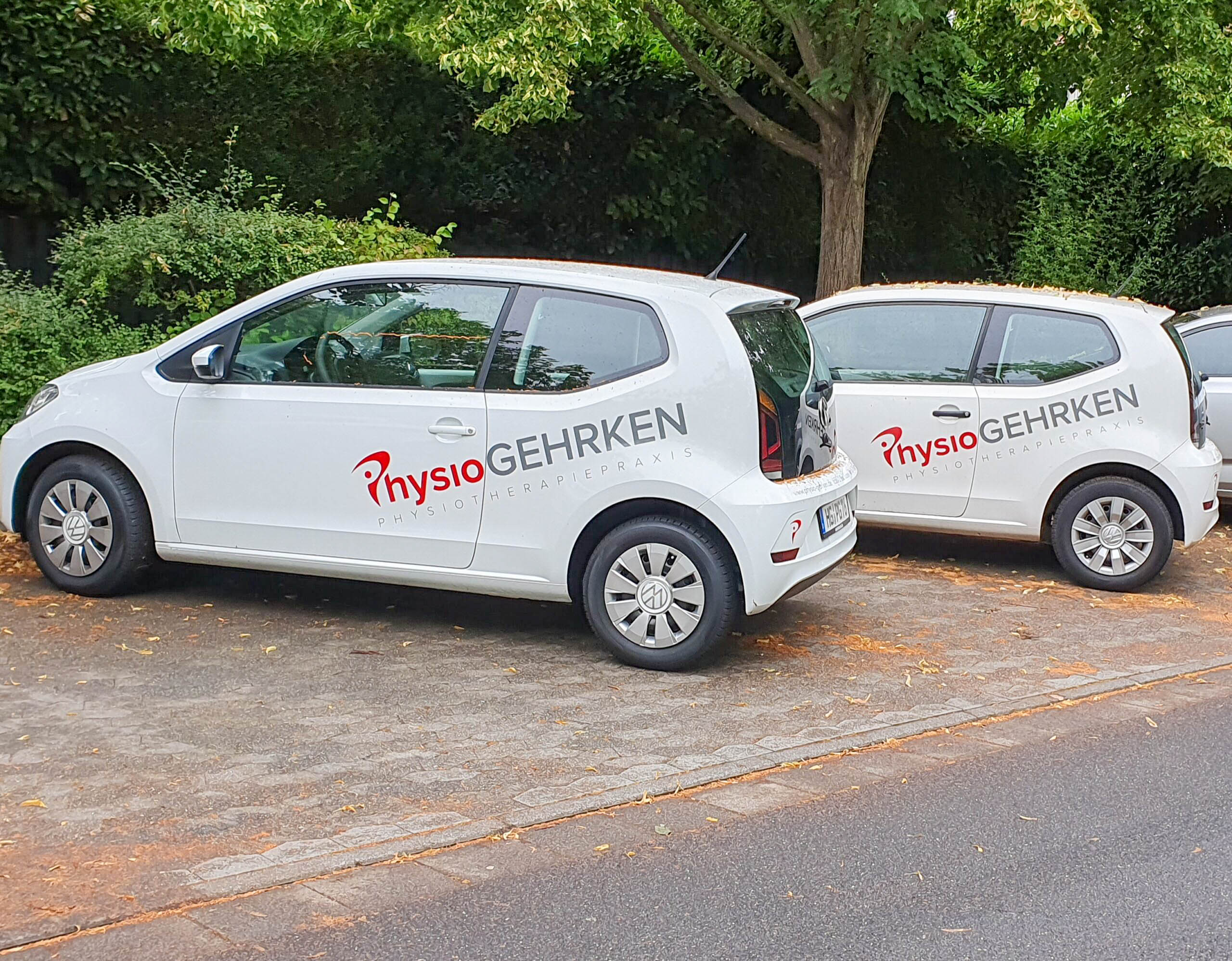 Zwei weiße Autos parken auf einer Straße, beide sind mit der Aufschrift „Physio Gehrken“ in roter und schwarzer Schrift versehen. Im Hintergrund sind Bäume und Laub zu sehen.