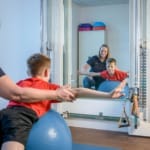 Ein Physiotherapeut hilft einem kleinen Jungen, der vor einem Spiegel auf einem Gymnastikball das Gleichgewicht hält.